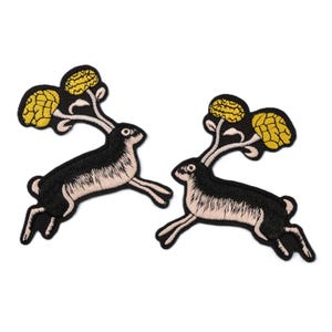 Könnte beinhalten: Zwei gestickte Aufnäher mit einem schwarzen Hasen mit cremefarbenen Details, der mit gelben Blumendesigns springt. Die Aufnäher haben eine detaillierte, strukturierte Oberfläche, ideal für die Stoffdekoration. Jeder Aufnäher ist etwa 7 cm lang.