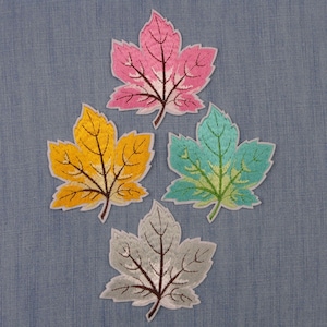 May include: Four embroidered leaf patches in shades of pink, yellow, teal, and gray. Each leaf has a white border and detailed stitching. The patches are arranged on a blue denim background, showcasing their vibrant colors and intricate designs.