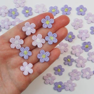 May include: A collection of small, embroidered flower appliques in shades of lavender, lilac, and white. Each flower features a yellow center. Some appliques are held in a hand, while others are scattered on a white surface.