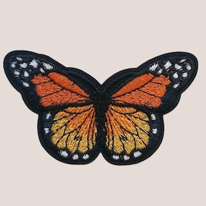 Pode incluir: Um remendo de borboleta bordado preto com asas laranja e amarelas. A borboleta tem um contorno preto e manchas brancas nas asas.