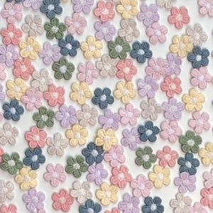 Puede incluir: Una colección de pequeñas aplicaciones de flores bordadas en varios colores pastel, incluyendo rosa, amarillo, azul, verde y morado. Las flores están dispersas sobre una superficie blanca, creando un patrón delicado y caprichoso. Ideal para manualidades y adornos.