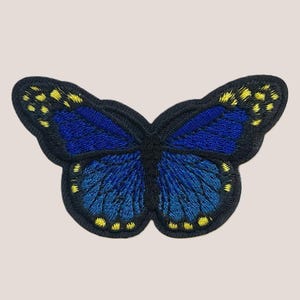 Pode incluir: Um remendo bordado com uma borboleta azul com detalhes amarelos. A borboleta tem contornos pretos e é um design detalhado.