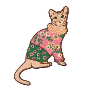 Peut inclure: Écusson brodé d'un chat assis portant un pull rose avec des motifs floraux et une jupe verte à pois blancs. Le chat a un col floral jaune et un corps brun clair avec des yeux verts.