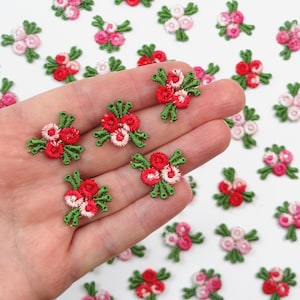 Op de afbeelding: Geborduurde bloemenapplicaties in rood, roze en wit met groene bladeren. Deze kleine, gedetailleerde bloemenpatches zijn verspreid over een wit oppervlak, sommige worden in een hand gehouden. Ideaal voor knutselen en versieringen.