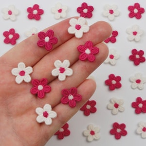 以下が含まれることがあります： 白とフクシア色の小さな刺繍の花のアップリケのクローズアップ。花は、対照的な中心部を持つ5つの花びらのデザインです。これらの装飾的なアップリケは、衣類、アクセサリー、またはクラフトプロジェクトを飾るのに最適です。