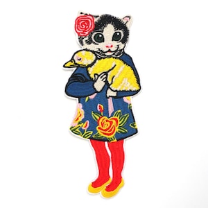 以下が含まれることがあります： 頭に赤い花をつけた白黒の顔の猫の刺繍ワッペン。猫は黄色い花柄の青いドレスを着て、黄色いアヒルの子を抱えています。このキャラクターは鮮やかな赤色のレギンスを着用しています。