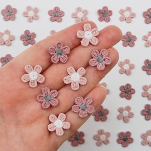 May include: A collection of small, embroidered flower appliques in shades of pink, mauve, and white. Each flower has a gray center and delicate stitching, perfect for crafting and embellishments.