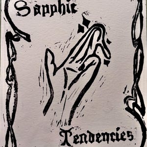 May include: Black and white linocut print on paper. The artwork features a stylized hand with the words "Sapphic Tendencies" in a gothic font. The image is framed by a decorative border.