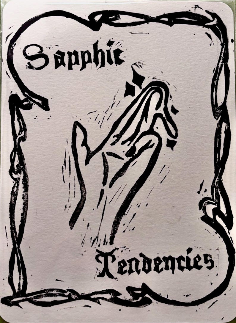 May include: Black and white linocut print on paper. The artwork features a stylized hand with the words "Sapphic Tendencies" in a gothic font. The image is framed by a decorative border.