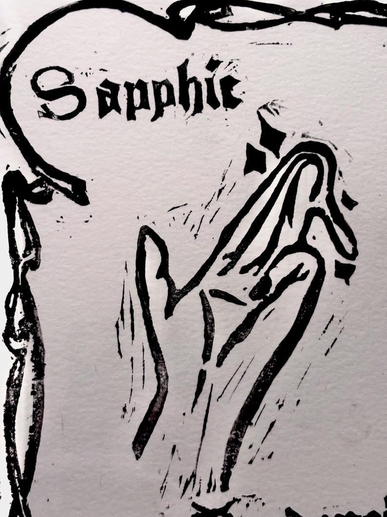 May include: A black and white linocut print with the word "Sapphic" in a gothic font. The print depicts a stylized hand and decorative border elements, all in bold black ink on a light background.