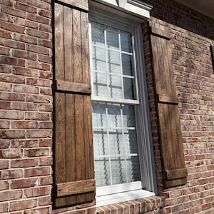May include: A window with white wooden shutters on a brick wall. The shutters are closed and have a dark brown stain.