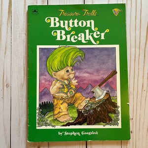 May include: A vintage children's book titled "Treasure Trolls Button Breaker" by Stephen Cosgrove. The cover is green with a colorful illustration of a troll with green hair and yellow clothing, holding an axe next to a tree stump.