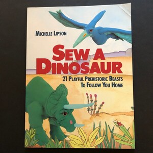May include: A book cover titled "Sew a Dinosaur" by Michelle Lipson. The cover features a green triceratops and a blue pterodactyl, with text that reads "21 Playful Prehistoric Beasts To Follow You Home."