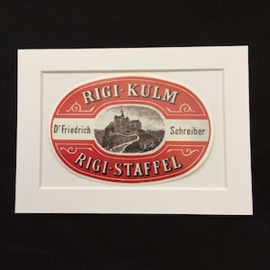 May include: A red oval label with white text that reads "Rigi-Kulm" and "Rigi-Staffel". The label features a black and white illustration of a building with a winding path leading up to it. The text "D' Friedrich" and "Schreiber" are also visible on the label.