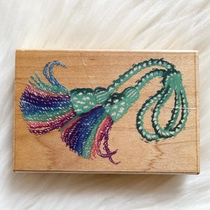 May include: A wooden stamp with a green and blue tassel design. The tassel is made of rope and has a fringe at the end.