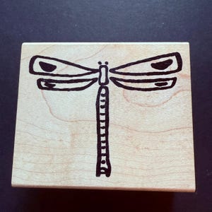 May include: A wooden stamp featuring a black dragonfly design. The dragonfly has detailed wings and a segmented body. The stamp is rectangular and made of light-colored wood, perfect for crafting and scrapbooking projects.
