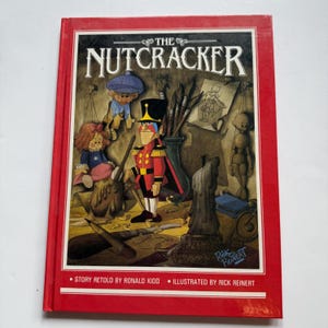 May include: A hardcover book titled "The Nutcracker" with a red cover. The cover illustration features a nutcracker soldier, dolls, and art supplies. Text includes "Story Retold by Ronald Kidd" and "Illustrated by Rick Reinert".