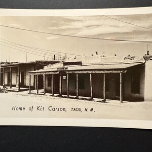 May include: Black and white photograph of a building with a covered porch supported by wooden posts. The text "Home of Kit Carson, TAOS, N.M." is printed at the bottom of the image. The building has a flat roof and appears to be made of adobe.