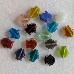 May include: A collection of small, translucent glass beads shaped like frogs in various colors, including red, blue, yellow, green, purple, black, brown, and clear. Each bead features a distinct frog silhouette with a raised ridge along the center.
