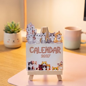 May include: A rectangular 2027 calendar card featuring colorful cartoon cat illustrations at the top and bottom, displayed on a small wooden easel. The word CALENDAR and the year 2027 are printed in the center in a brown, rounded font style.
