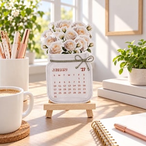 May include: A desktop calendar shaped like a mason jar filled with pale pink roses. The calendar displays the month of January 2027 with a grid of dates. It is supported by a small wooden easel stand. The design features a decorative twine bow.