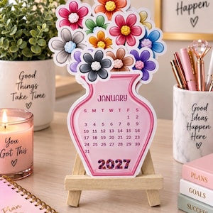 May include: A die-cut desk calendar shaped like a pink vase filled with colorful flowers. The calendar displays the month of January 2027 with a grid of dates. It is supported by a small wooden easel stand. The design features various floral colors.