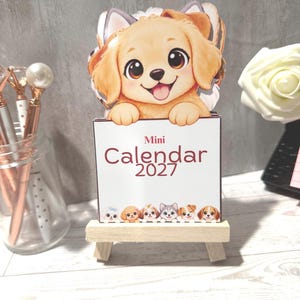 May include: A mini 2027 calendar featuring a cartoon golden puppy illustration at the top and a row of various small dog breeds at the bottom. The calendar is displayed on a small, light-colored wooden easel stand, showcasing the text Mini Calendar 2027.