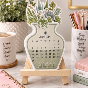 May include: A die-cut desk calendar shaped like a vase filled with various green and sage-colored succulents. The calendar displays the month of January 2027 with black text on a light green background, resting on a small, light-colored wooden easel.