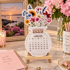 May include: A 2027 January calendar shaped like a vase filled with colorful illustrations of sunflowers, lilies, and lavender. The calendar is displayed on a small wooden easel and features the text 2027 at the top, with the month January and the days of the month listed below in a clear, black font.