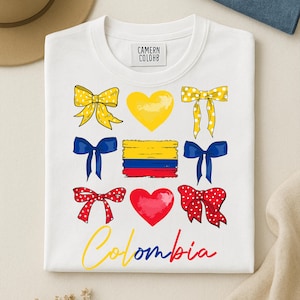 May include: White crew neck sweatshirt with a colorful graphic design. The design includes yellow bows, a yellow heart, blue bows, the Colombian flag, red bows, a red heart, and the word "Colombia" in yellow and red.