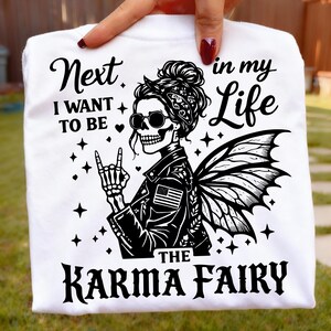 May include: White t-shirt with a black graphic of a skeleton fairy wearing sunglasses and a leather jacket with an American flag patch. Text reads "Next I Want to Be in my Life The Karma Fairy."