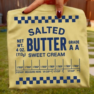 May include: A pale yellow t-shirt featuring a blue graphic design that mimics a butter package. Text includes "SALTED BUTTER," "NET WT. 113g," and measurement guides. The shirt is held up by a person.