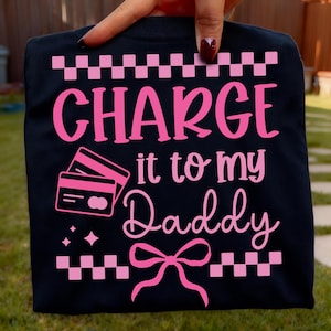 May include: Black t-shirt with the phrase "CHARGE it to my Daddy" in pink, accompanied by pink credit card graphics, stars, and a bow. The top and bottom of the shirt have a pink and white checkered pattern.