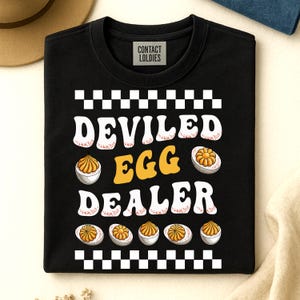 May include: Black sweatshirt with the text "DEVILED EGG DEALER" in white and yellow, with images of deviled eggs. The top and bottom of the design have a black and white checkered pattern. The brand "CONTACT LOLDIES" is printed on a small white label.