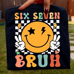May include: Black sweatshirt with a retro smiley face design. The design includes an orange smiley face with star eyes and lightning bolt accents, peace signs, and the words "SIX SEVEN BRUH" in colorful, bubble letters.