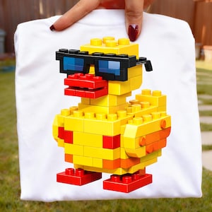 May include: White t-shirt with a graphic of a yellow duck constructed from toy bricks. The duck wears black sunglasses with blue lenses and has a red beak and feet. The design is centrally placed on the shirt.