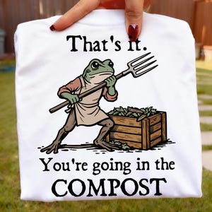 May include: White t-shirt with a humorous graphic of a frog holding a pitchfork, standing near a wooden compost bin. The text reads "That's it. You're going in the COMPOST."