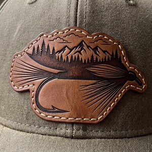 May include: A brown leather patch with a fly fishing lure design. The patch features a detailed mountain and forest scene above the lure, with white stitching around the edge. The patch is attached to a green hat.