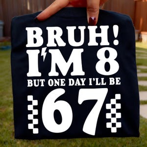 Peut inclure: T-shirt noir avec texte blanc : « BRUH ! J'AI 8 ANS, MAIS UN JOUR J'AURAI 67 ANS ». Le chiffre 8 est en grand format. Le chiffre 67 a un motif à carreaux sur les côtés.