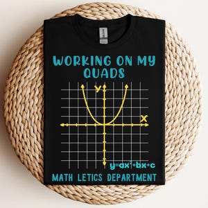 May include: Black t-shirt with the text "WORKING ON MY QUADS" in turquoise, and a yellow parabola graph. Below the graph is the equation "y=ax1+bx+c" and the words "MATH LETICS DEPARTMENT". The shirt is displayed on a woven, circular surface.