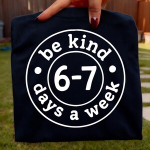 Może przedstawiać: Granatowa koszulka z białym okrągłym nadrukiem. Na nadruku napis "be kind 6-7 days a week". Koszulka trzymana jest przez dłoń z czerwonym lakierem do paznokci.