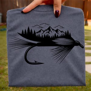May include: A gray t-shirt featuring a black graphic of a fly fishing lure with a mountain and forest scene above it. The design includes a hook, feathers, and a detailed mountain range with trees and clouds.