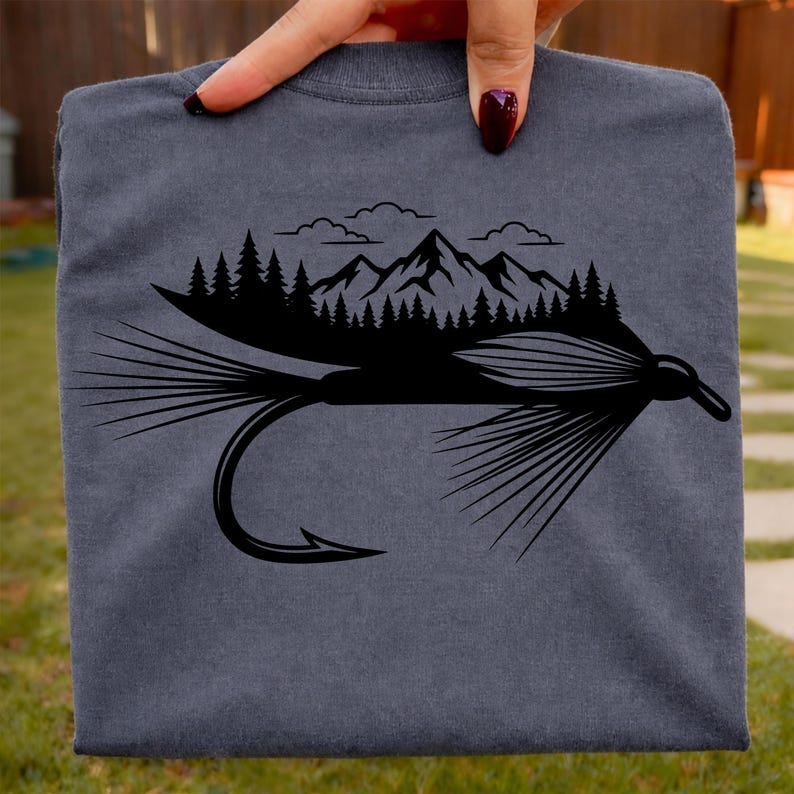May include: A gray t-shirt featuring a black graphic of a fly fishing lure with a mountain and forest scene above it. The design includes a hook, feathers, and a detailed mountain range with trees and clouds.