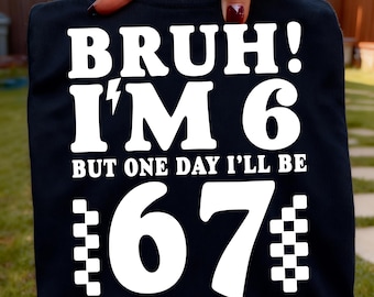 May include: Black t-shirt with white text that reads "BRUH! I'M 6 BUT ONE DAY I'LL BE 67". The number 67 is styled with a checkered pattern on the sides. The shirt is held up by a hand.