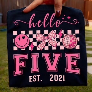 Puede incluir: Camiseta negra con un diseño gráfico rosa. El diseño incluye la palabra "hello", una carita sonriente, un lazo, una bola de discoteca y la palabra "FIVE". También se incluye el texto "EST. 2021". El diseño tiene un patrón de cuadros.