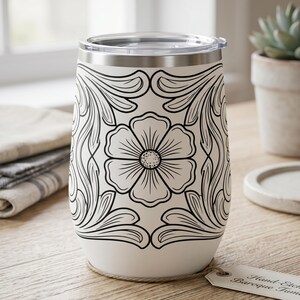 May include: A white stainless steel tumbler with a black line art floral pattern featuring a central flower surrounded by symmetrical scrolling leaves. The tumbler has a clear plastic lid and a metallic rim, with a small paper tag attached at the base.