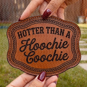 May include: A brown leatherette coaster with the phrase "HOTTER THAN A Hoochie Coochie" in black script. The coaster has a decorative border and is held by a person.