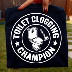 Op de afbeelding: Zwart T-shirt met een witte afbeelding. De afbeelding is een cirkelvormig ontwerp met de woorden "TOILET CLOGGING CHAMPION" rond een tekening van een toilet en een ontstopper. Twee sterren staan aan weerszijden van het ontwerp.