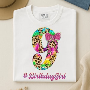 Op de afbeelding: Wit T-shirt met een groot nummer 9. Het nummer combineert luipaardprint en tie-dye patronen, met een roze glitterstrik. De tekst "#BirthdayGirl" staat eronder in roze glitter.