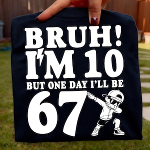 May include: Black t-shirt with white text that reads "BRUH! I'M 10 BUT ONE DAY I'LL BE 67". A cartoon illustration of a boy dabbing is on the bottom right. The shirt is held up by a hand.
